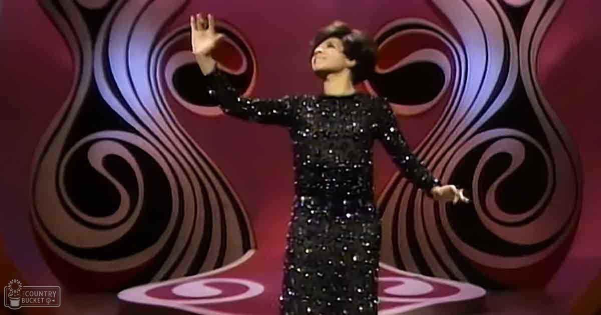 An adult female performer in a long-sleeved black sequined dress on stage with a stylized red, black, white, and pink backdrop.