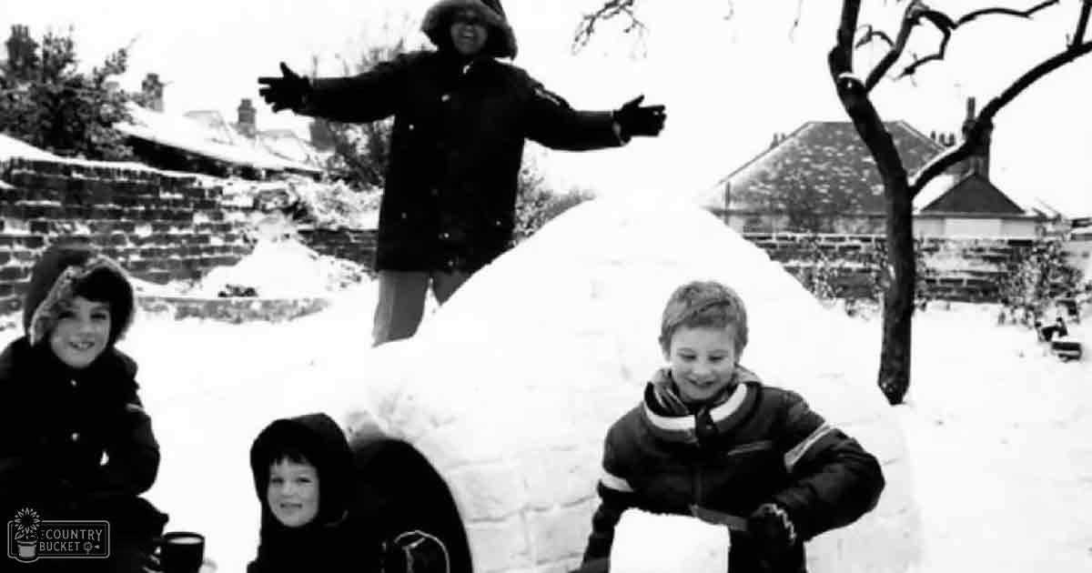 Children playing in snow with an adult; winter outdoor residential setting with bare trees and snow-covered ground.