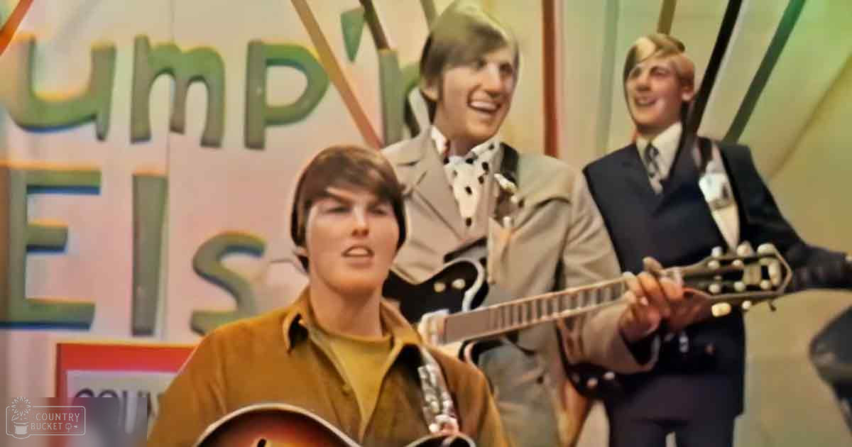 A musical performance scene with three young adult men holding acoustic guitars, set indoors under bright stage lighting.