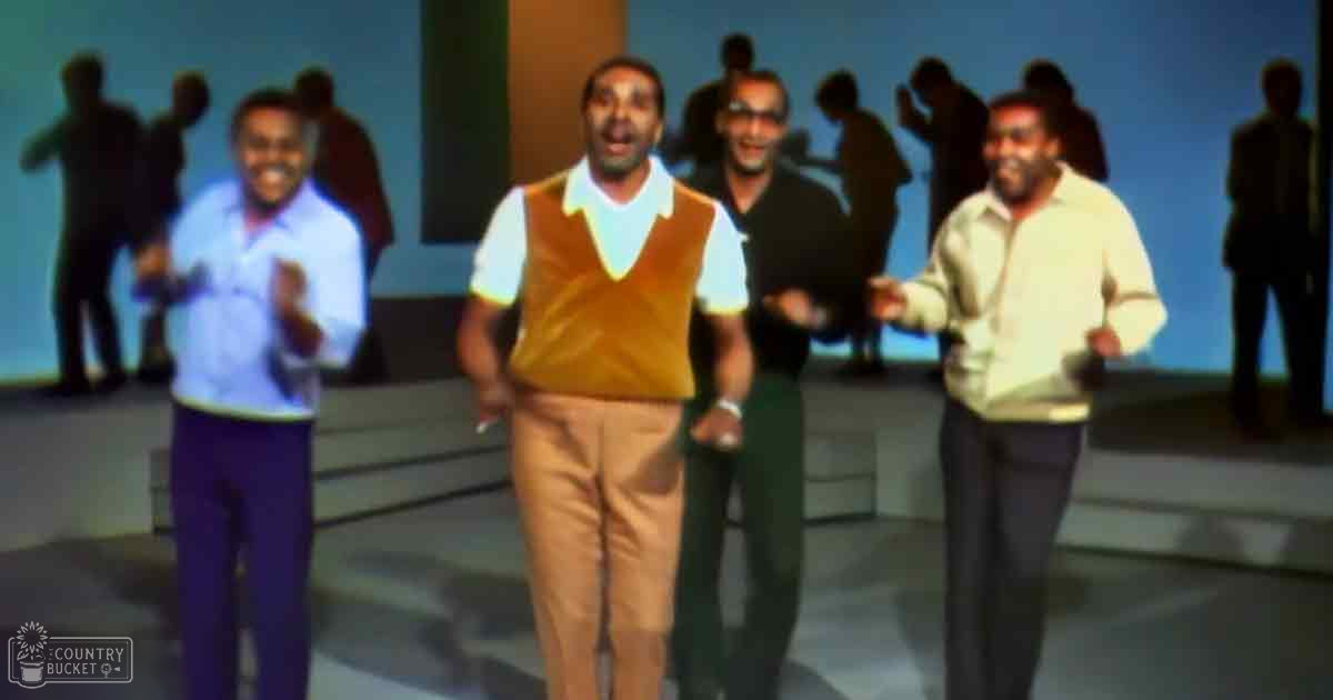 An adult male singer in a white shirt, mustard-colored vest, beige pants, and dark shoes stands center stage at an event or performance. Two other men smile on either side, all wearing casual attire.