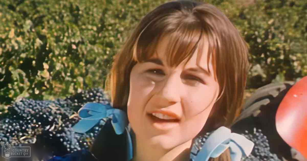 A young adult female wearing a flowy, dark blue polka-dot dress with puffed sleeves stands outdoors among greenery. A red balloon is partially visible in the frame.