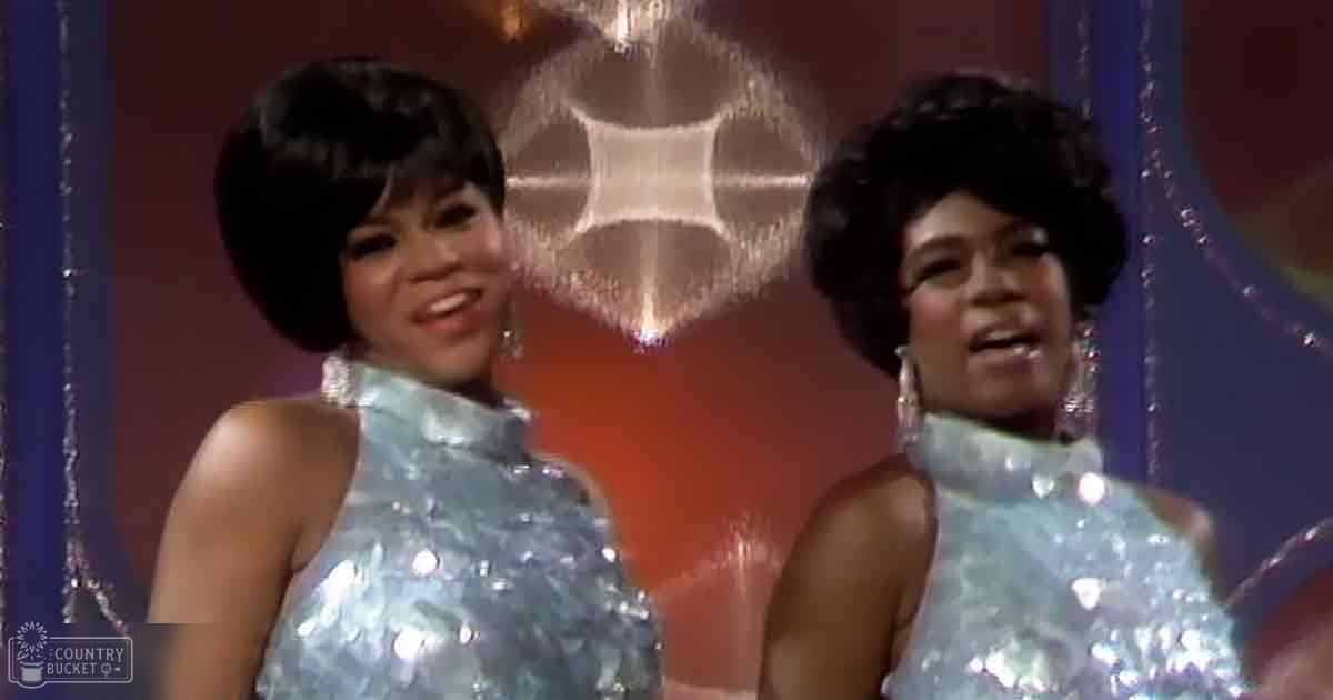 Two adult female performers in a stage setting, wearing sleeveless halter-neck sequined dresses with a silvery-blue or teal color, standing on a stage with dark curtains and a chandelier.