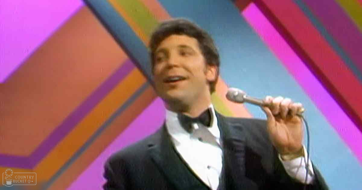 Adult male performer wearing a formal tuxedo jacket and bow tie holding a microphone against a vibrant geometric background, likely during an evening stage show.