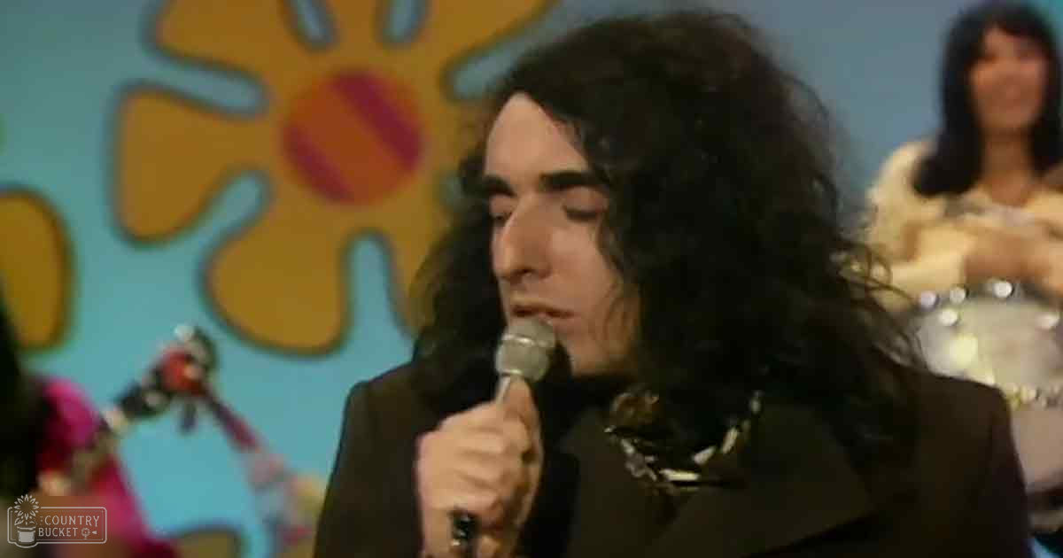 An adult male performer singing into a microphone at an indoor music event, surrounded by colorful floral decorations and another individual in the background holding what appears to be a tambourine.