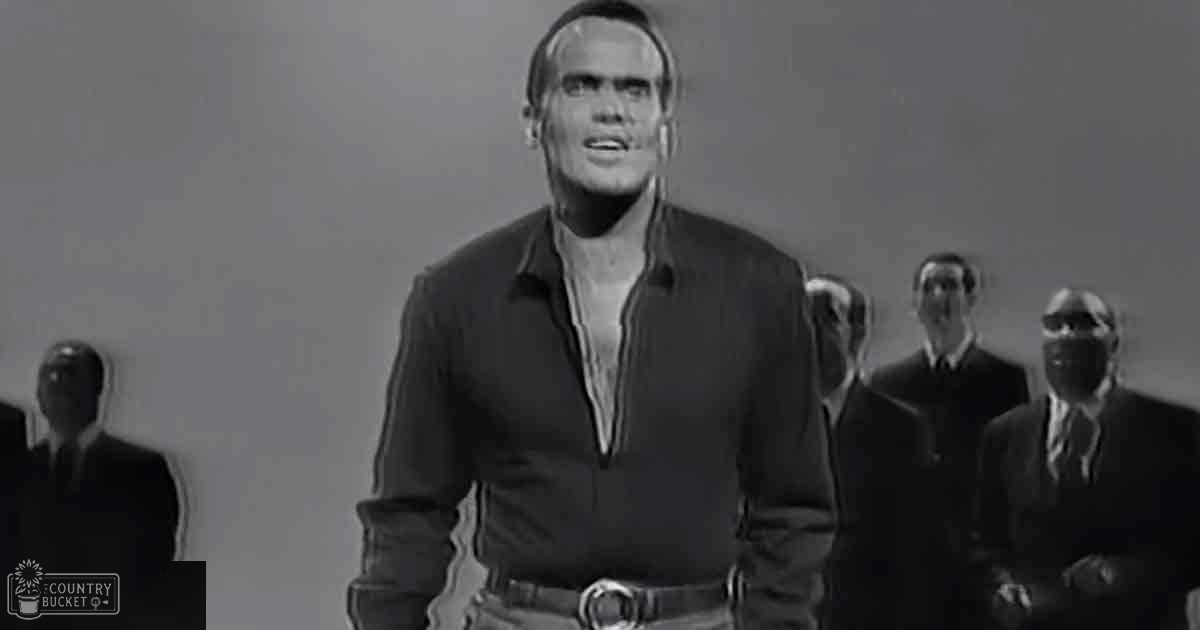 An adult male singer stands prominently at the center of a simple indoor setting with varying ages of men in formal attire surrounding him, suggesting a studio or stage environment.