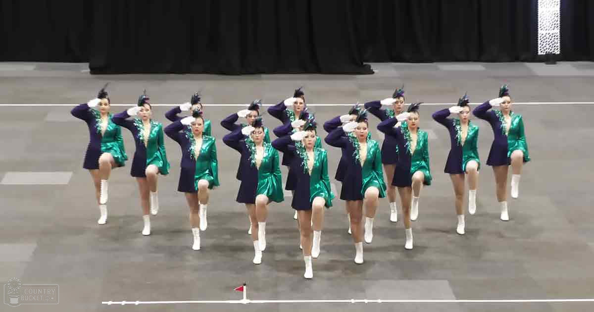 A synchronized dance group of young adult females performing on a dark stage with a black curtain backdrop, wearing shiny green jackets with decorative elements and white ankle-high boots under indoor lighting.