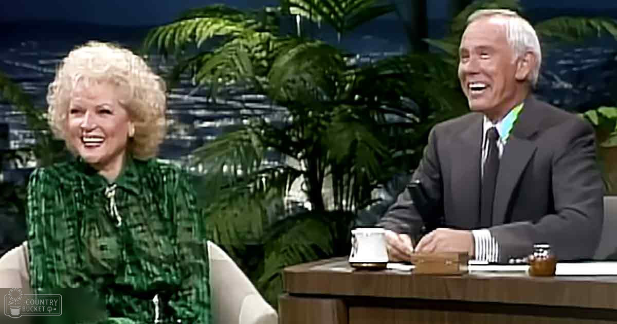 Talk show set with two older adults seated at a desk, with green patterned blouse and dark suit jacket in an indoor studio setting with large potted plants and cityscape backdrop.