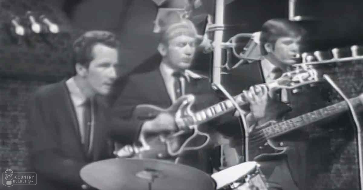 A musical performance with three adult males in a professional recording studio setting, with guitars and drums.