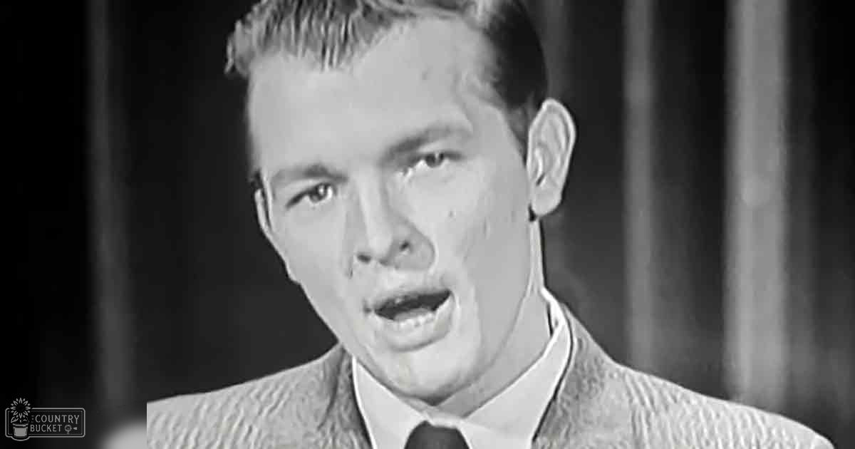 Young adult male in a fitted suit jacket with a textured pattern, standing indoors against dark curtains under focused lighting.