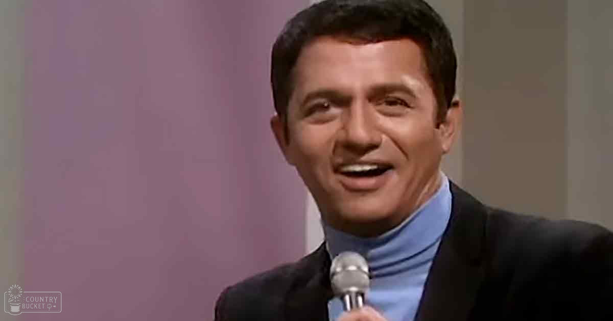 An adult male wearing a dark suit jacket and light blue turtleneck, standing indoors with a microphone held close to his mouth, suggesting he is speaking or performing.