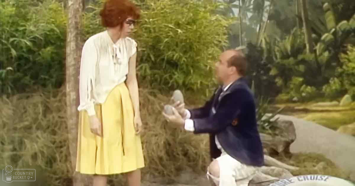 An adult male proposes marriage to an adult female in a lush tropical outdoor setting with palm trees, sign reading 'Cruise' visible.