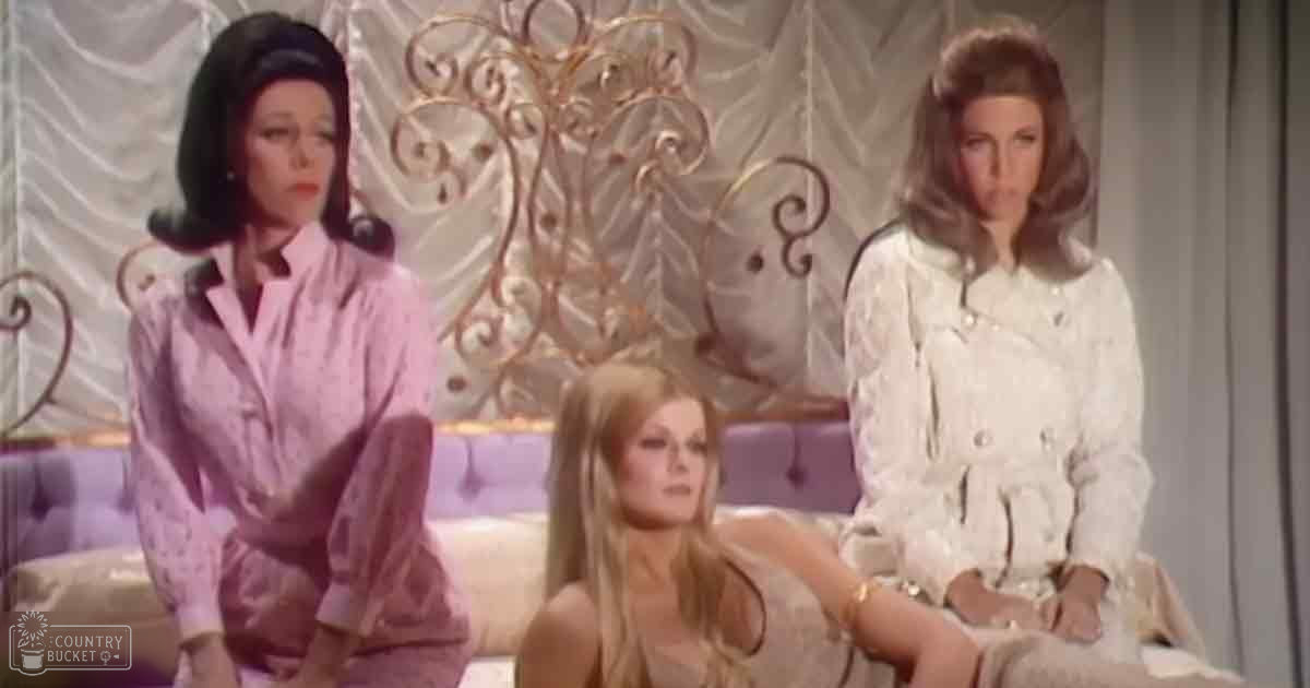 A cozy bedroom scene with three adult females dressed in light-colored loungewear; a bed with ornate gold headboard, sheer curtains, and light-colored bedding, set against soft indoor lighting.