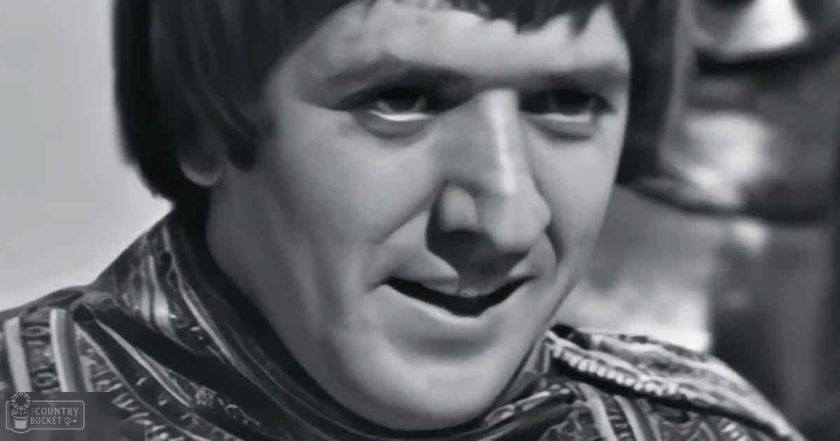 A middle-aged adult male in a Roman-style tunic or armor with intricate patterns, set indoors with minimal background details.