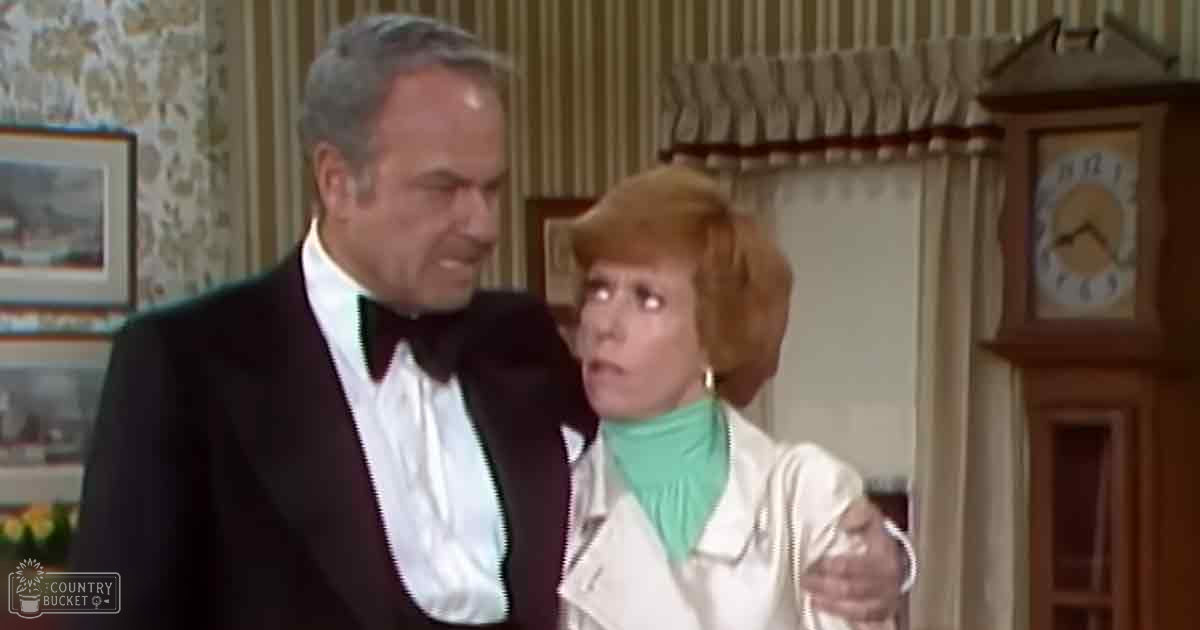 A middle-aged man in a formal black tuxedo with white shirt and bow tie stands indoors alongside a woman wearing a white jacket over a green turtleneck, both from an earlier era. A wooden grandfather clock is visible on the right.