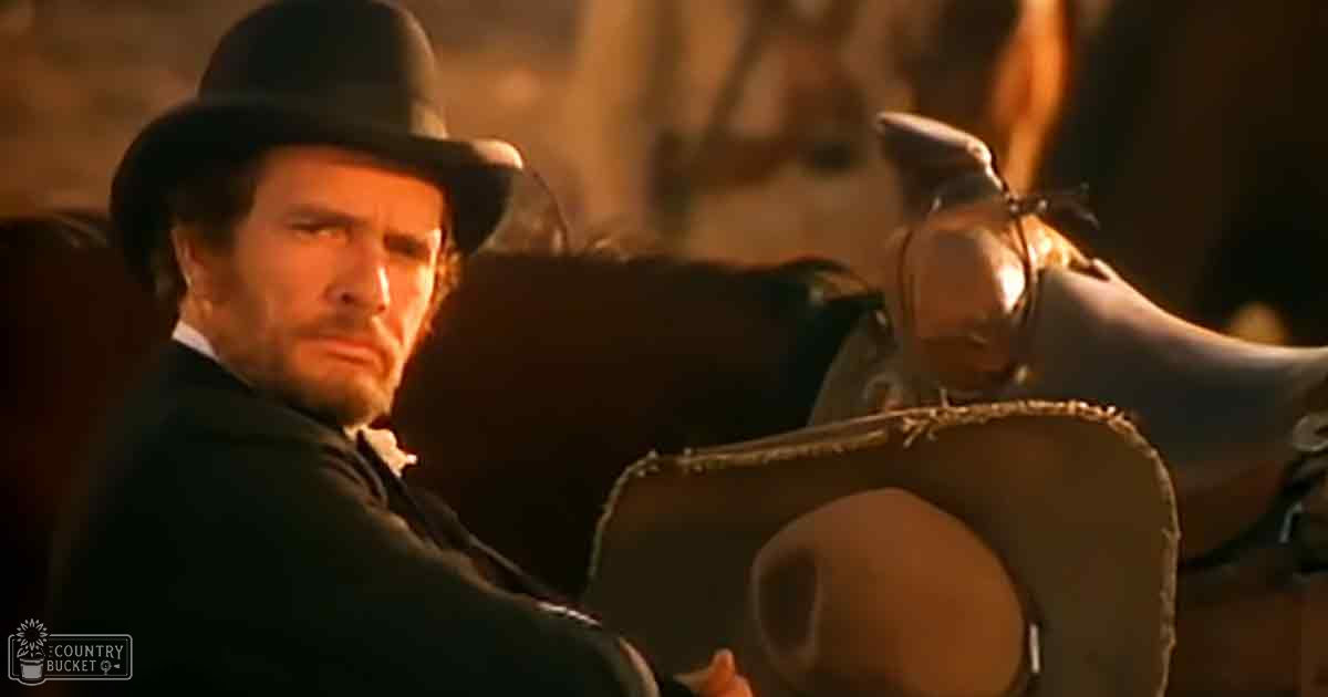 An adult male in a dark suit and white shirt with a black hat, interacting with a dark brown horse in an indoor Western setting with rustic elements and warm lighting suggesting evening or nighttime.