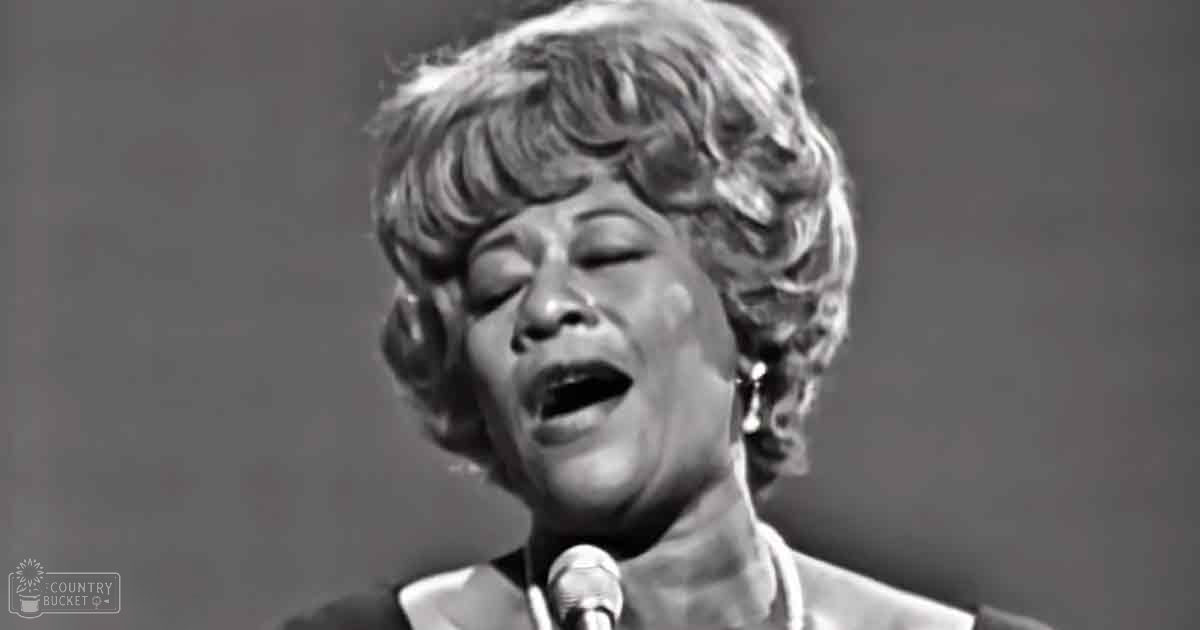 A middle-aged female singer in a dark-colored mid-20th-century style garment sings into a microphone against a plain backdrop, suggesting an indoor performance or recording session.