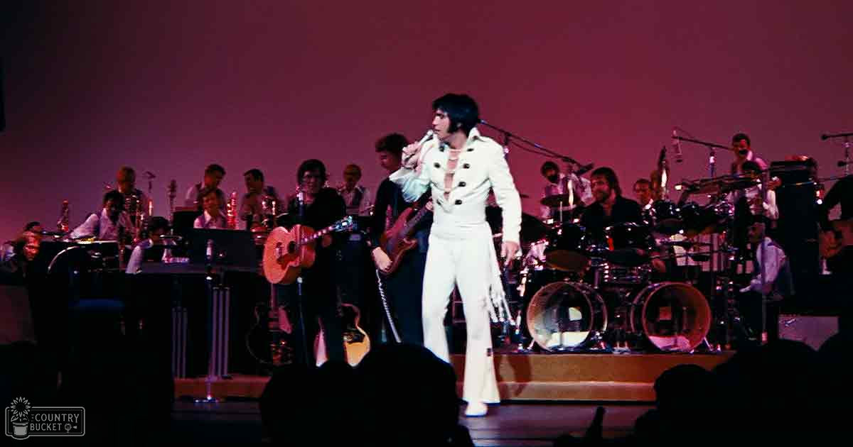 An adult male performer in a white double-breasted suit with fringes at the bottom of the pants, likely from a musical performance setting.
