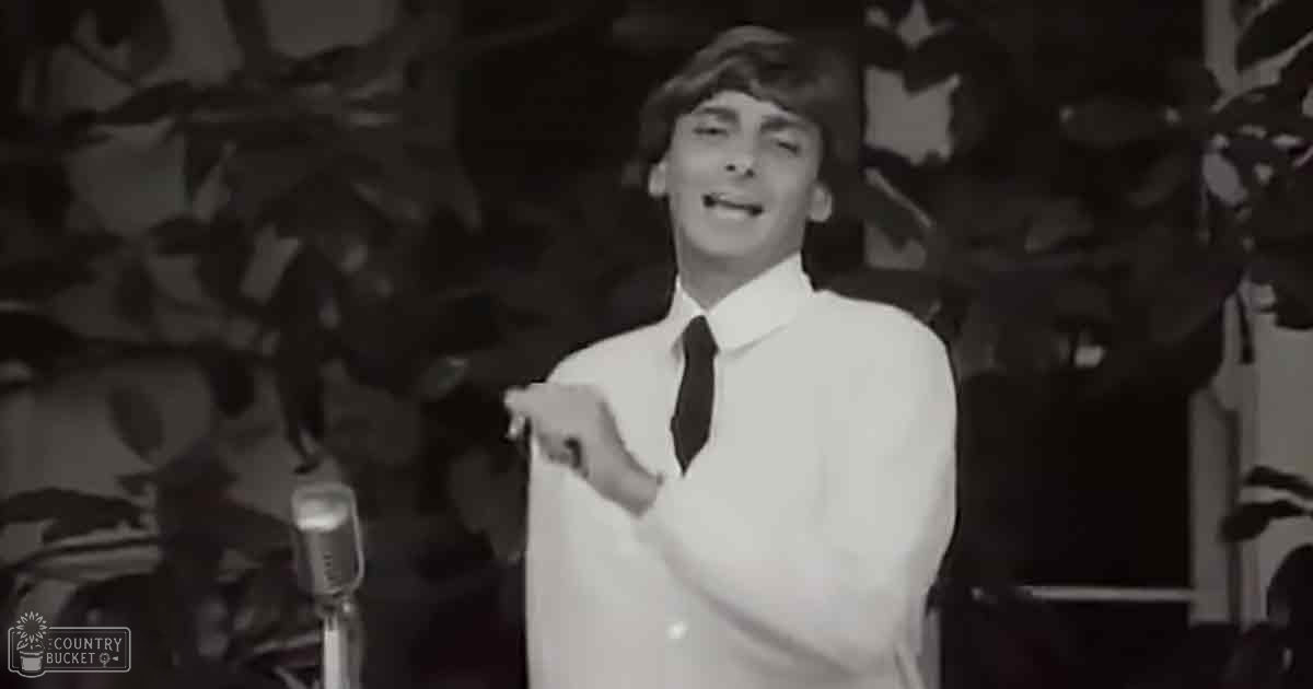 A young adult male in a performance setting, wearing a white long-sleeved shirt and dark tie, with plants or foliage behind him suggesting an indoor stage environment.