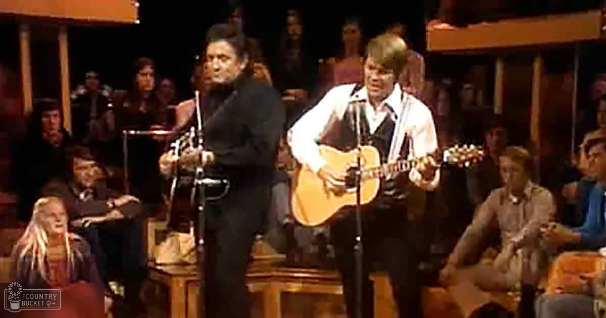 An adult male singer in a black suit jacket and white shirt, accompanied by another young adult male holding an acoustic guitar, performing in front of an audience seated in rows of chairs with wooden furniture, likely at a concert hall or theater during the evening under artificial lighting.
