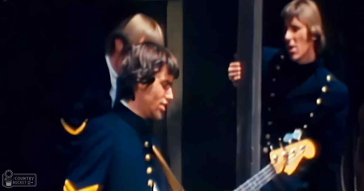 A musical performance scene with young adult males in dark blue military-style uniforms against a backdrop of indoor stage lighting.