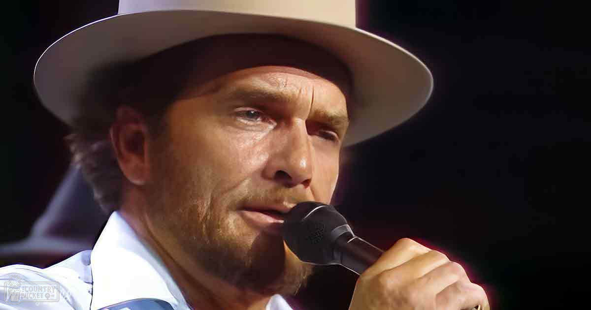 A middle-aged man wears a white shirt and wide-brimmed hat, holding a microphone against an indoor stage-like backdrop with focused lighting.