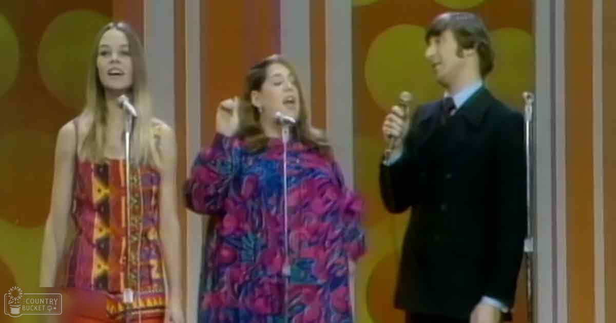 A musical performance scene with young adults wearing vibrant, patterned dresses and a man in a dark suit on stage against a colorful backdrop.
