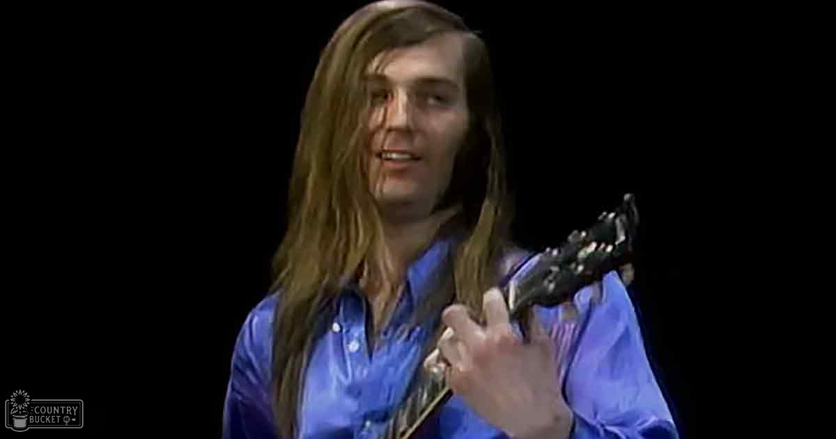 A young adult male playing a black electric guitar indoors, wearing a blue button-up shirt with the text 'Madly Odd' on it.