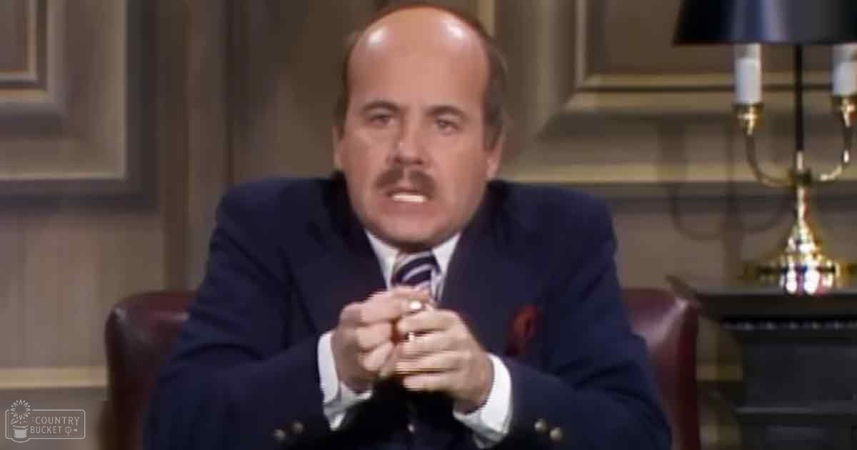 An older adult male wearing a dark blue suit jacket with a white shirt and patterned tie sits in an indoor setting with paneling and natural light.