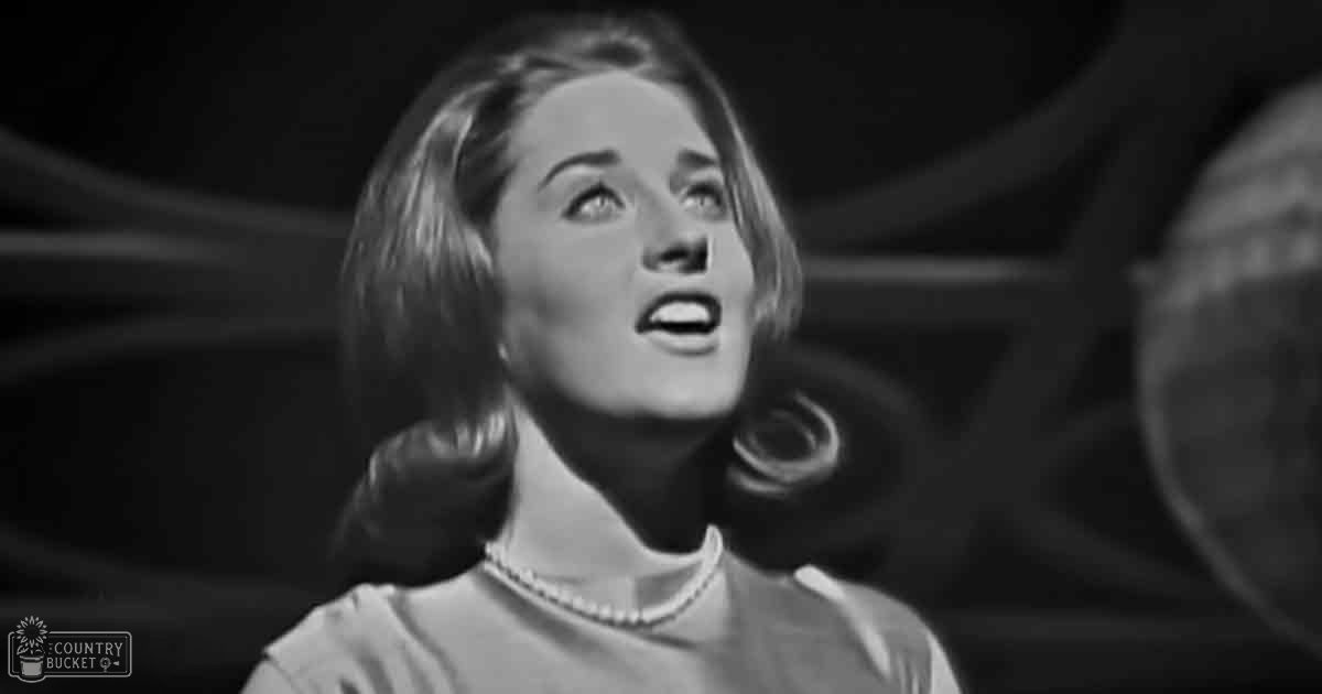 A young adult female wearing a light-colored, relaxed, collared blouse with short sleeves and a pearl necklace stands indoors against dark stage lighting, suggesting a theatrical or cinematic setting.