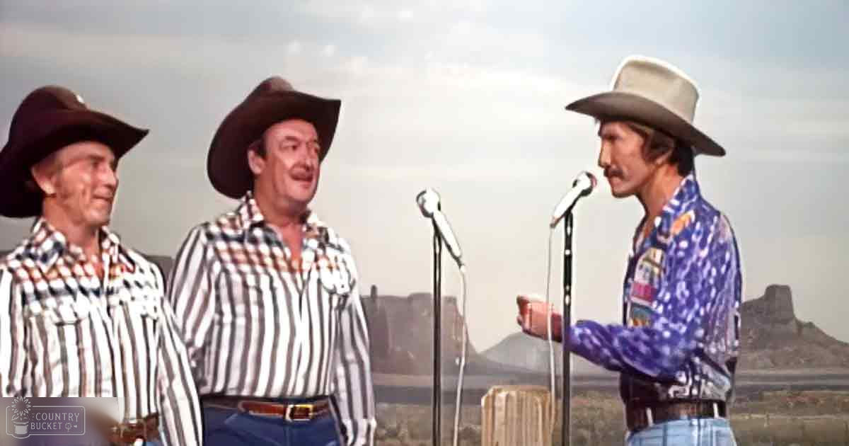 Three adult males wearing plaid shirts with cowboy hats perform outdoors against a desert backdrop under slightly overcast skies.