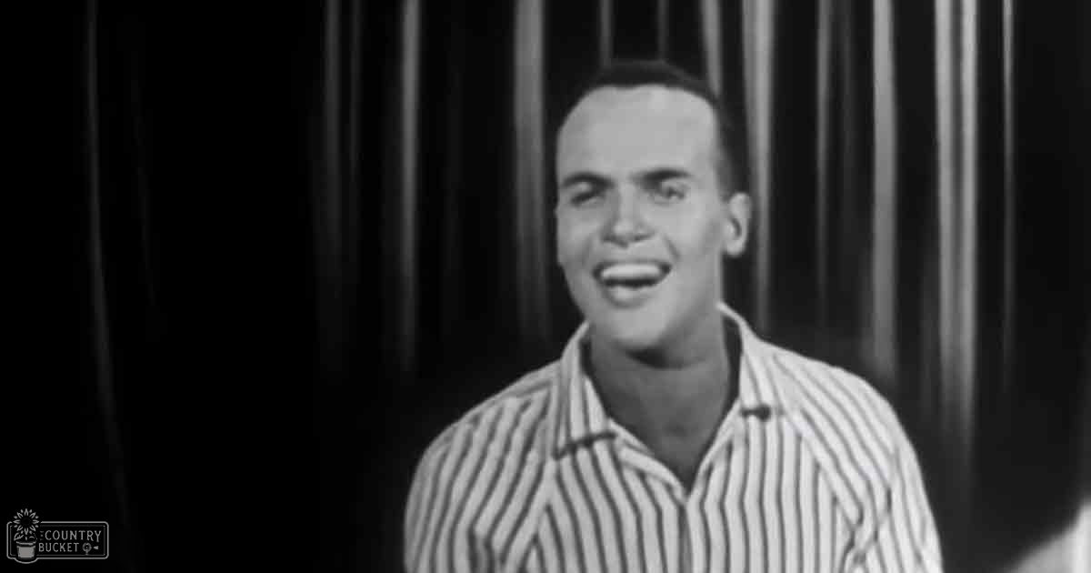 A young adult male in a striped shirt stands indoors with vertical blinds as the backdrop.