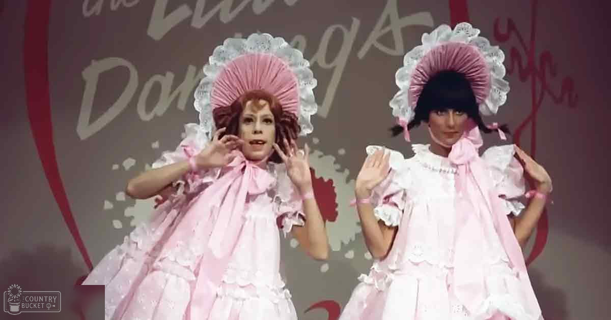 A theatrical performance scene with young adult female performers in vintage-style white and pink dresses, set indoors on a stage with red and white cursive text backdrop.