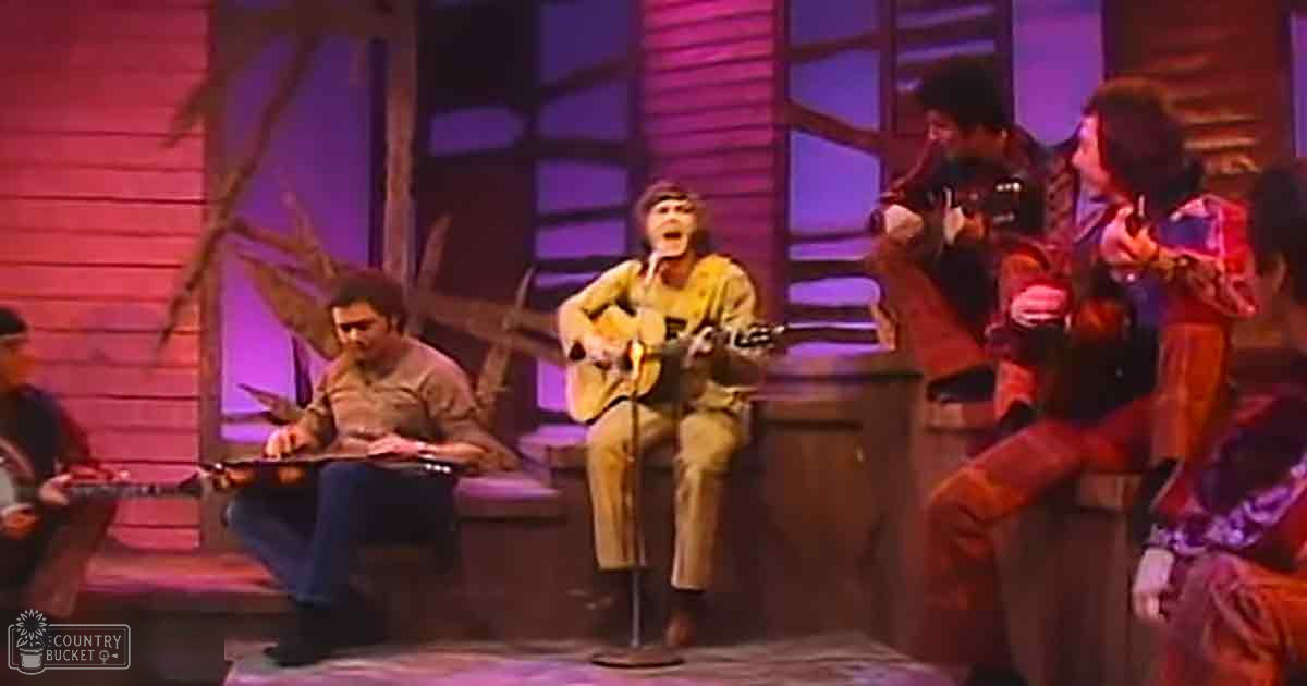 A lively indoor musical performance with a young male guitarist, an older male on guitar, and a female holding percussion in front of a stage with rustic props under warm artificial lighting.