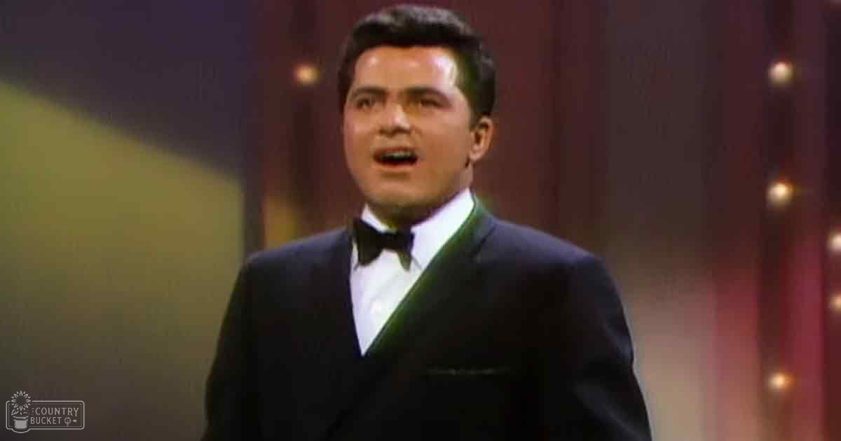 A male adult in a dark suit with a white shirt and black bow tie, standing on an indoor stage-like setting with curtains and stage lights.