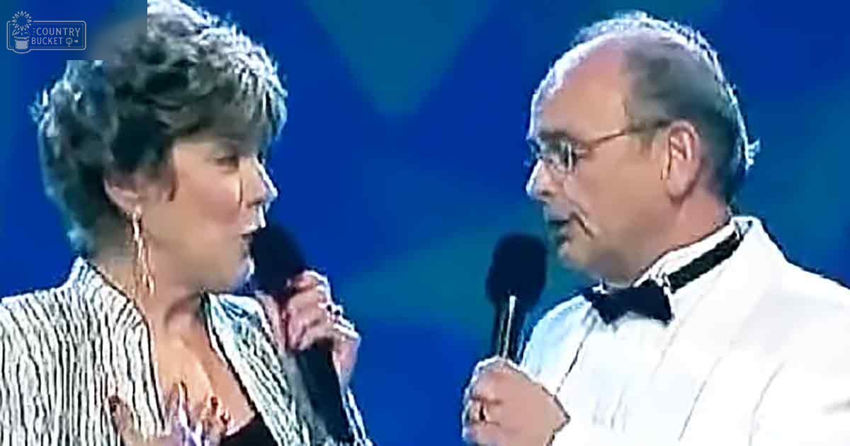 A middle-aged female singer and male performer stand side by side, each holding a microphone against a blue-lit stage backdrop. The female wears a striped blazer over a black top, while the male dons a white tuxedo jacket with a bow tie.