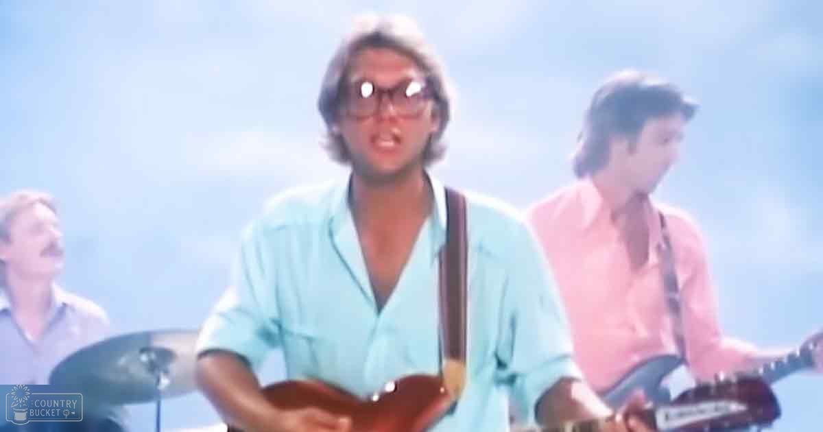 Young adult male playing guitar outdoors with a relaxed atmosphere, surrounded by other musicians.