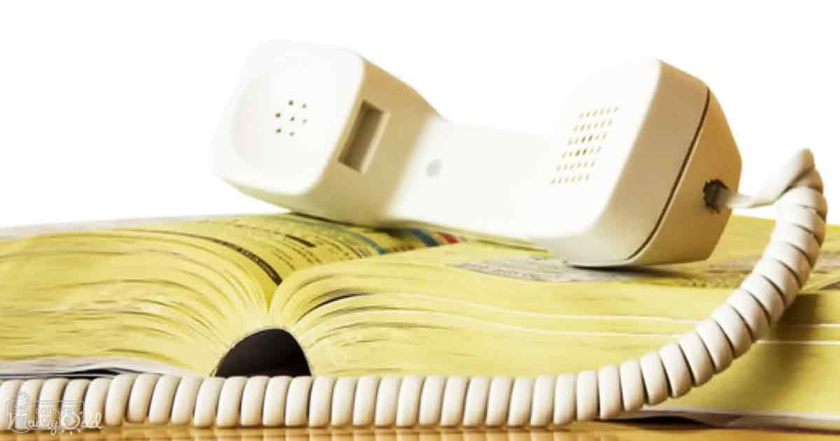 A white telephone handset with a coiled cord rests on an open book in an indoor setting, with plain white background.