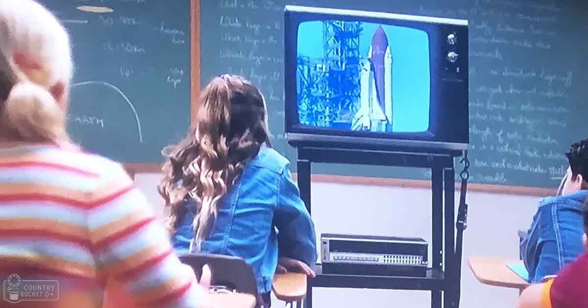 A classroom scene with children watching an older-style television displaying a space shuttle launch, wooden desks, chairs, and a blackboard filled with text.