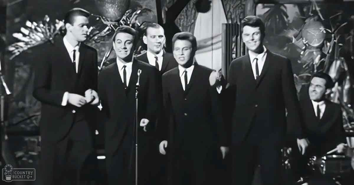 A mid-20th-century musical performance group dressed uniformly in dark suits, white shirts, black ties, and dark shoes stands on a stage with decorative foliage behind them. The setting suggests an indoor theater or concert hall during a live performance.