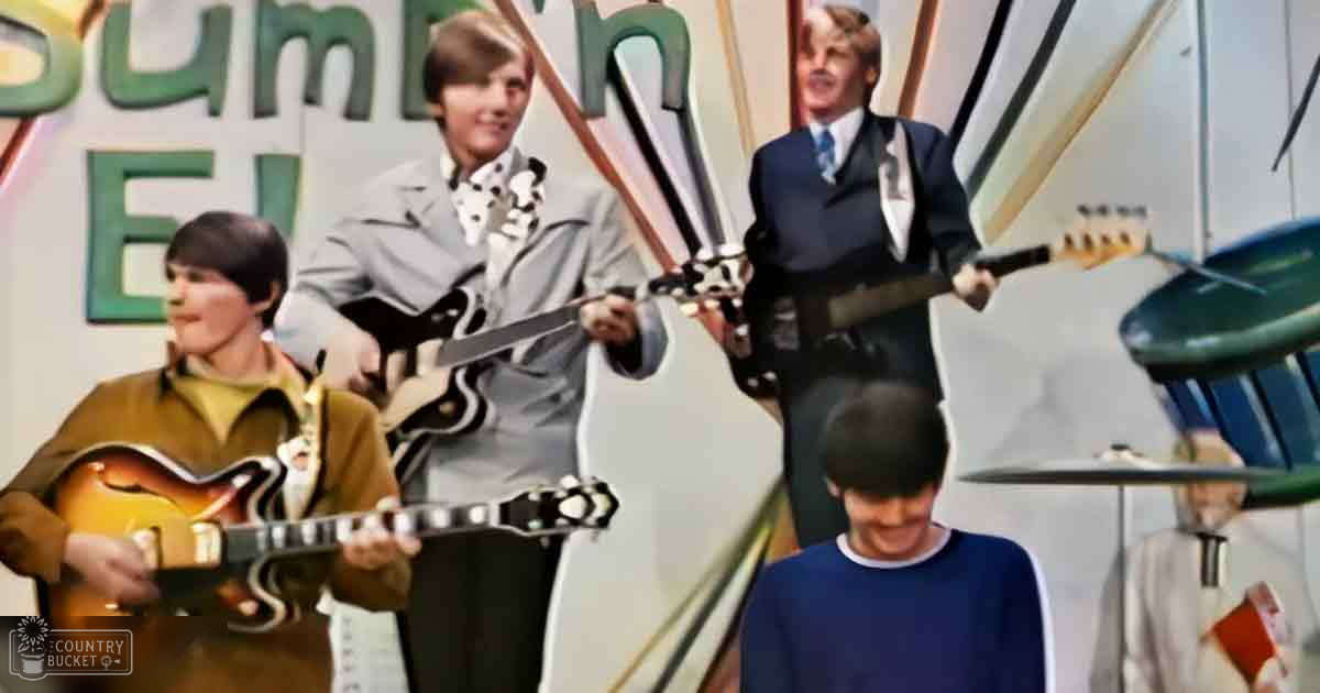 A group of musicians performing in front of a colorful, summer-themed backdrop with bright lighting and geometric designs.