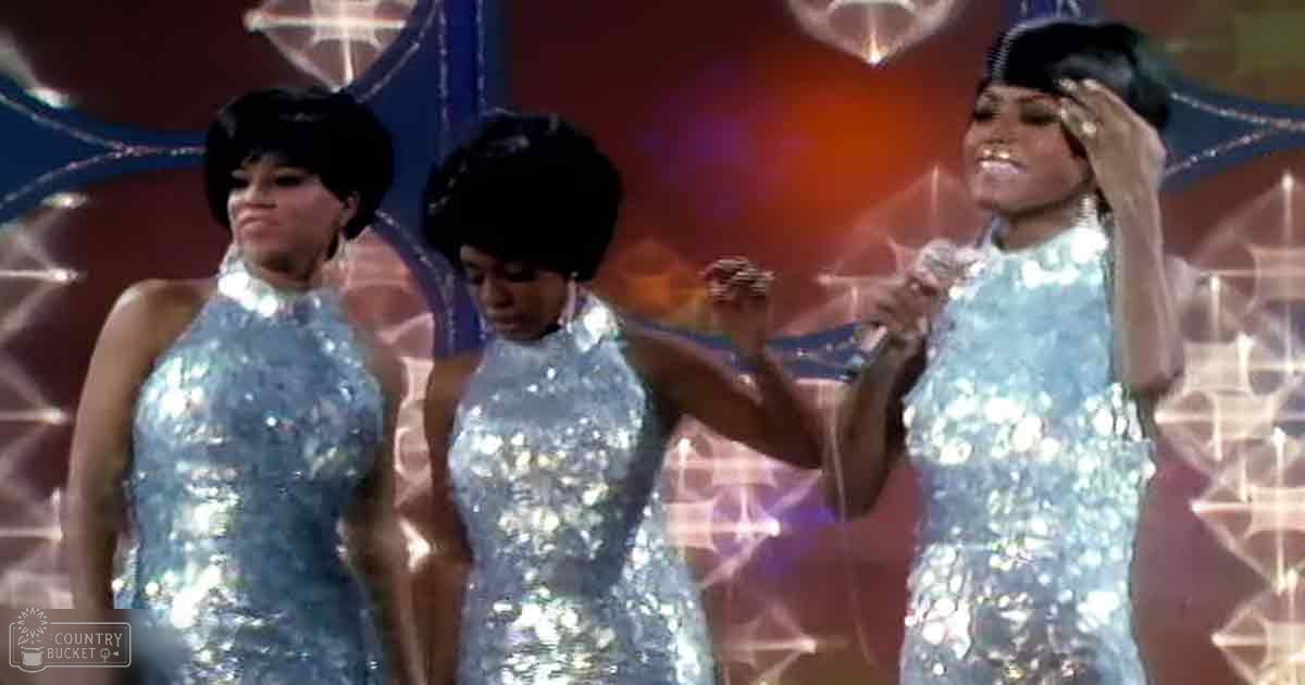 Three adult female performers in matching sleeveless high-necked sequined dresses on a stage with decorative lighting effects with star-shaped lights and red-blue hues.