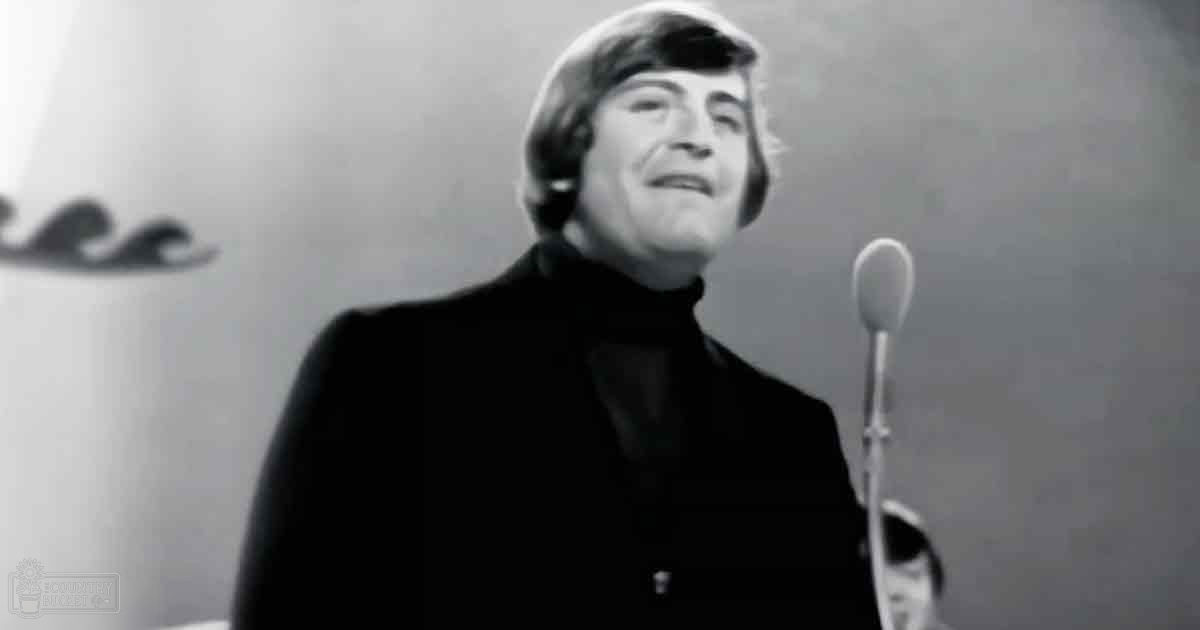 An adult male singer wearing a dark-colored jacket with a high collar and turtleneck, performing in an indoor setting under professional lighting.