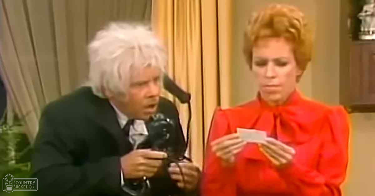A comedic scene from a television show set indoors with a male performer in a dark green suit and a female guest in a bright red dress holding a piece of paper, surrounded by furniture such as a chair and a wooden cabinet.