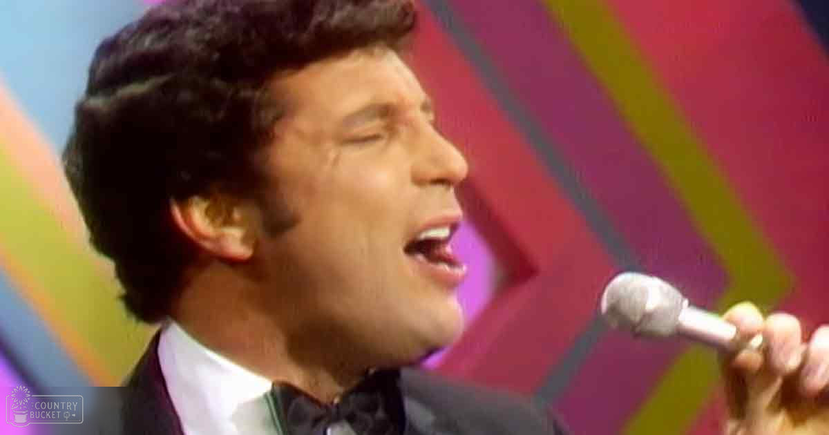 A male adult singer in a dark suit with white shirt and black bow tie, performing into a microphone against a vibrant stage backdrop from the 1960s or 1970s era.