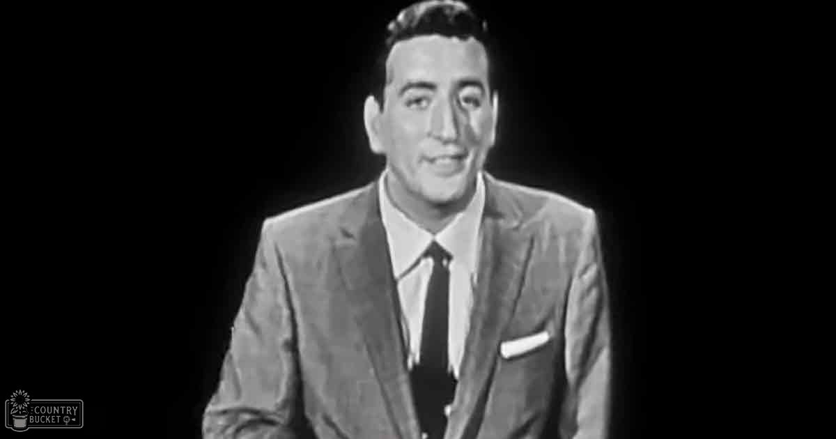 Young adult male in formal attire stands against dark backdrop, likely studio or stage setting; monochromatic photograph with dominant black and white tones.
