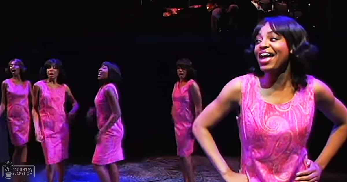Young adult female performer in a theatrical setting, wearing a sleeveless pink dress with patterned design and high neckline. Background performers also dressed similarly on stage under dramatic lighting.