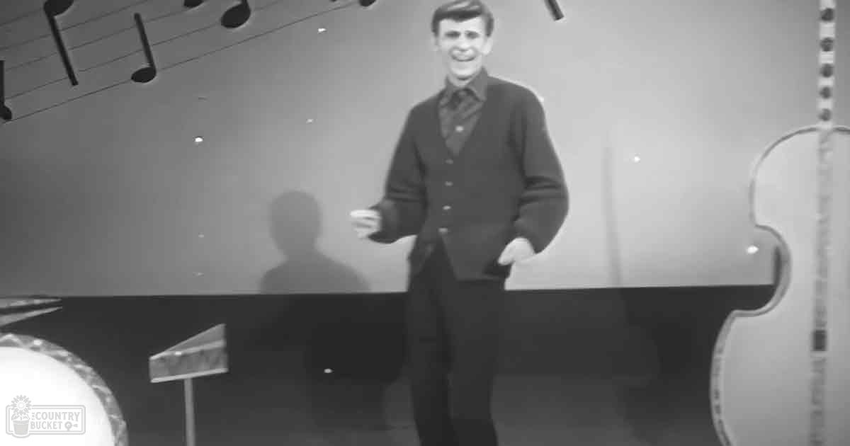 A young adult male dressed casually in a studio setting with musical instruments and notes on the wall.