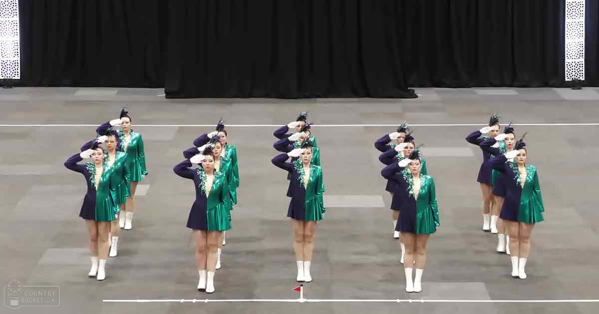 Young adult female dancers performing in an indoor arena or gymnasium, wearing shiny dark blue jackets with green accents and white trim around the collar and cuffs, paired with short shiny green skirts that end above the knee, white socks, and white boots.