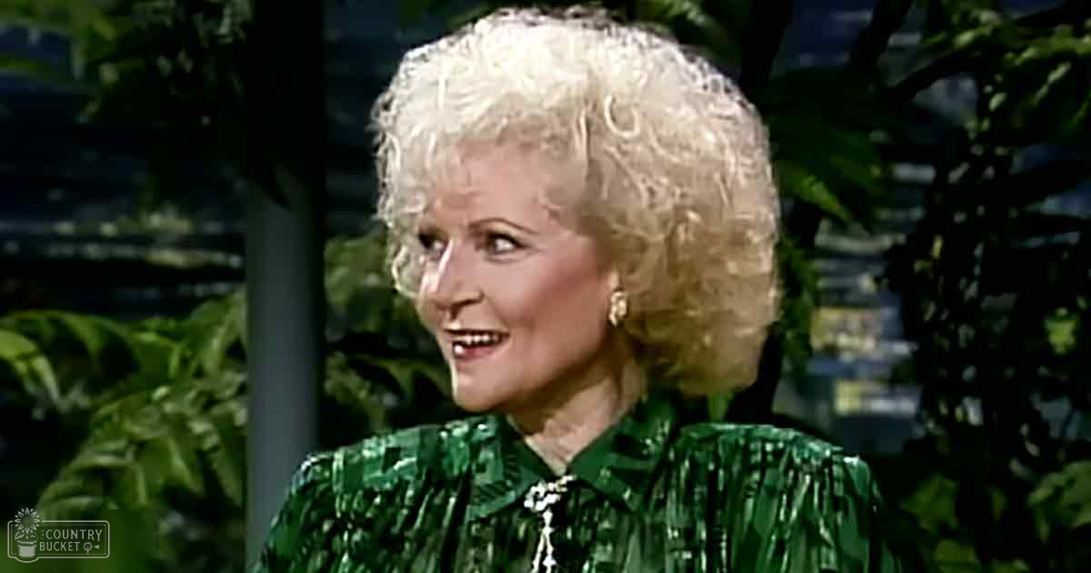 An older adult female in a garden setting, wearing a green, ruffled, long-sleeved blouse with gold hoop earrings.