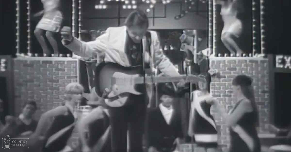A young adult male playing guitar on stage with an audience seated in the foreground; setting suggests a mid-20th century musical performance, possibly from the 1960s.